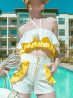 Woman standing in the summer sun wearing a white and yellow ruffled crop top and white shorts with yellow ruffles around the pockets