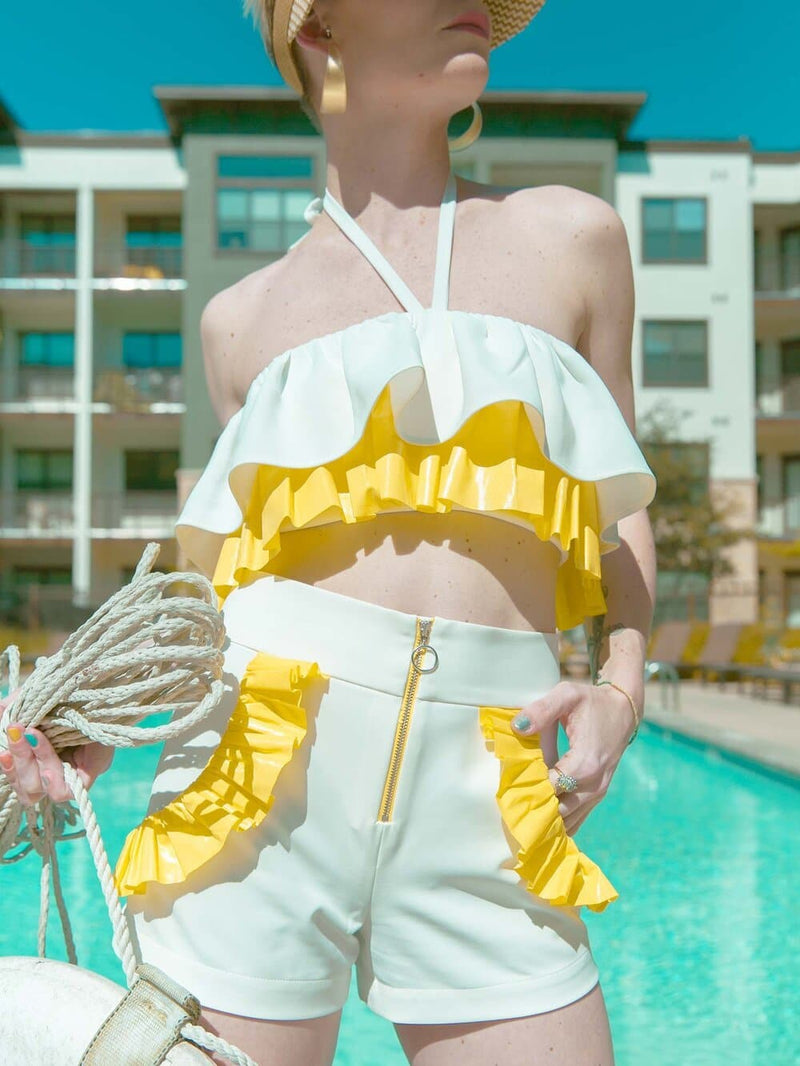 Woman standing nearby a pool wearing a white and yellow ruffled crop top and white shorts with yellow ruffles around the pockets
