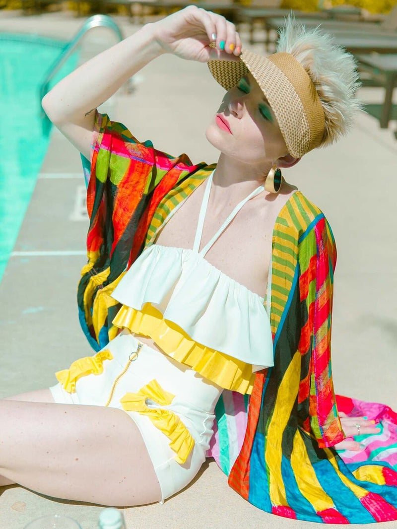 Woman sitting in the sun wearing a white and yellow summer crop top and shorts with yellow ruffles arouns the pockets