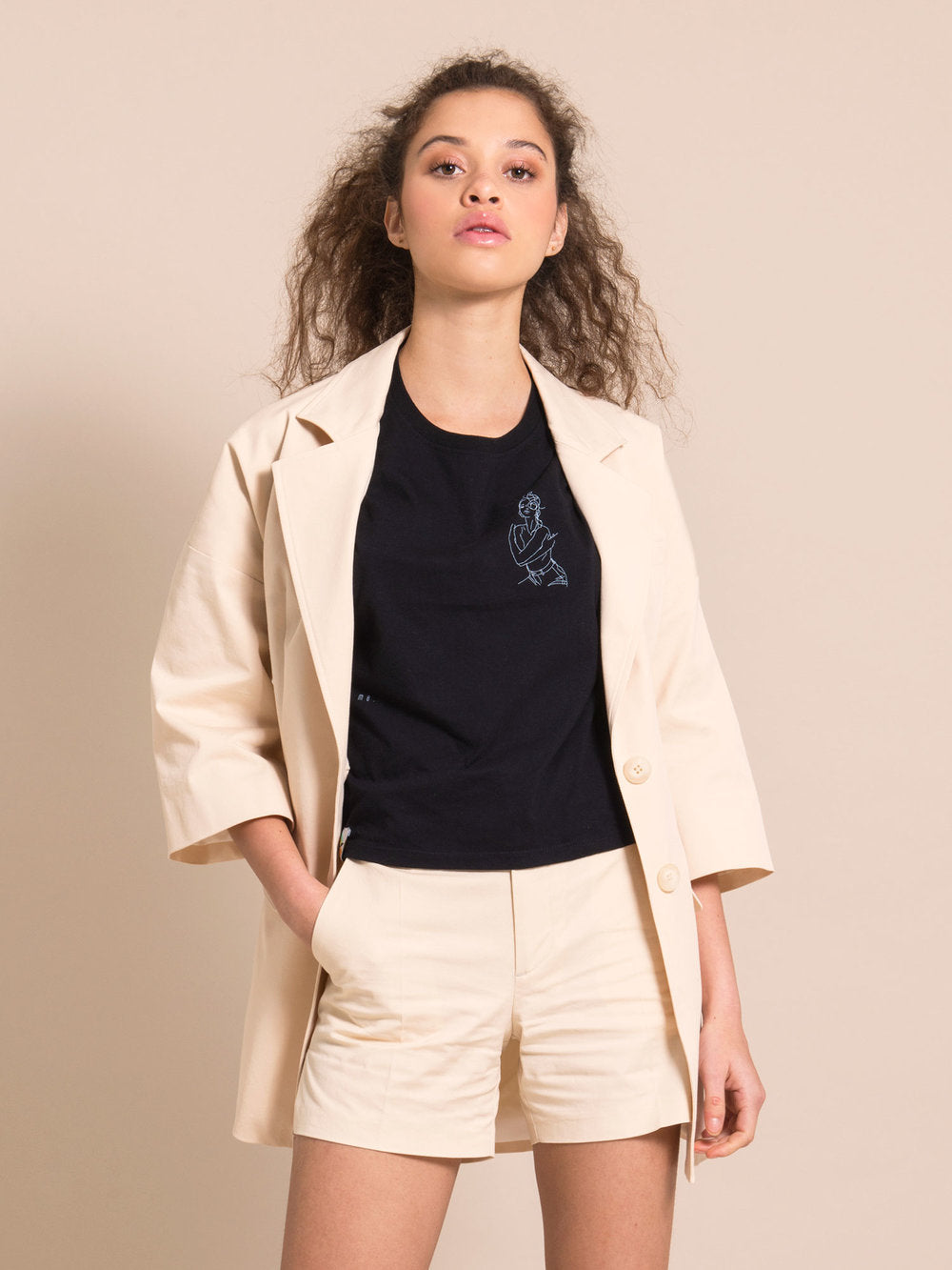 Woman wearing sustainable beige shorts with crease, black tee with print and a long beige blazer with bell sleeves